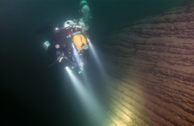 Subacqueo in configurazione tecnica illumina una parete stratificata nel Lago di Como durante un’immersione profonda