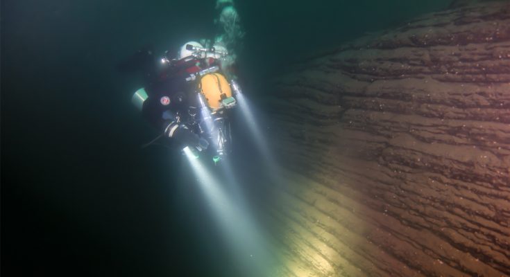 Subacqueo in configurazione tecnica illumina una parete stratificata nel Lago di Como durante un’immersione profonda