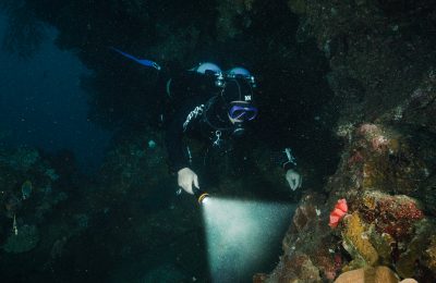 Subacqueo con attrezzatura tecnica esplora un relitto con torcia durante un'immersione in acque profonde.