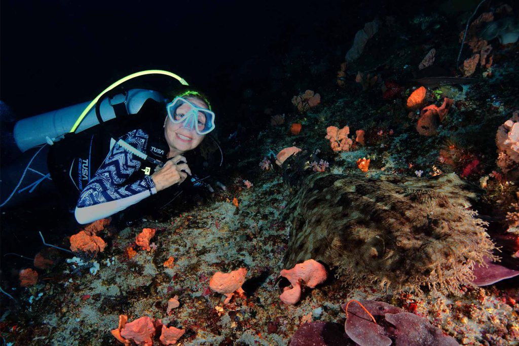 La subacquea Marina sorridente in immersione notturna osserva da vicino uno squalo wobbegong sul fondale corallino - Raja Ampat.