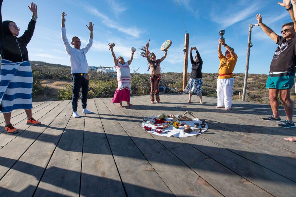 Gruppo di persone che partecipano a una cerimonia rituale all'aperto, con le braccia alzate verso il cielo. Un uomo vestito con abiti tradizionali regge un tamburo al centro, mentre gli altri sono disposti su una piattaforma di legno intorno a un'offerta floreale sul terreno, a Socorro, Messico.