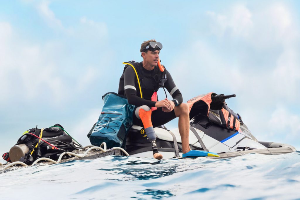 Subacqueo con protesi seduto su una moto d'acqua con attrezzatura da immersione e zaino impermeabile Groundtruth x PADI.
