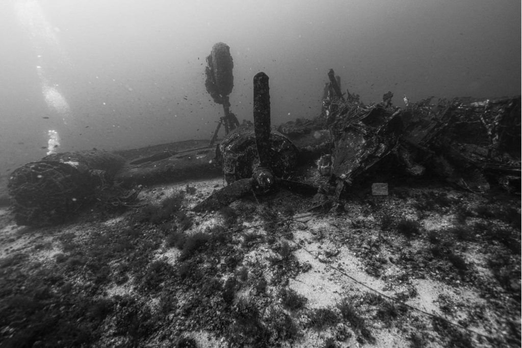 Vista in bianco e nero del relitto di un B-24 Liberator sul fondale marino, con motore ed eliche parzialmente sepolti nella sabbia