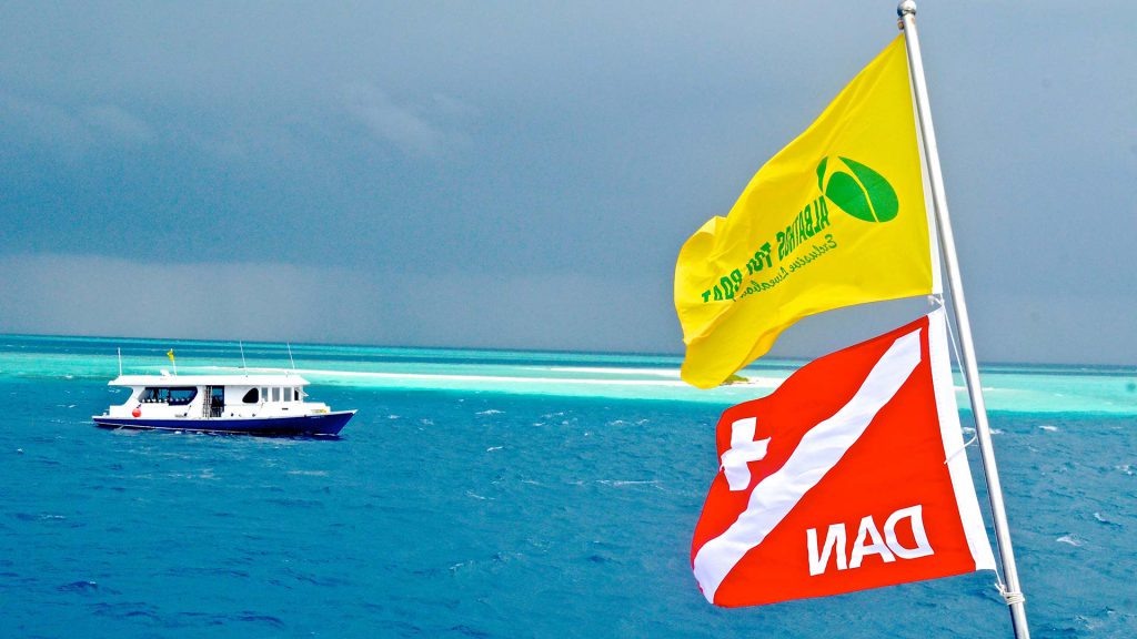 Bandiere Albatros Top Boat e DAN sventolano in navigazione alle Maldive, con dhoni sullo sfondo e reef degli atolli centrali crociera natale maldive subacquea