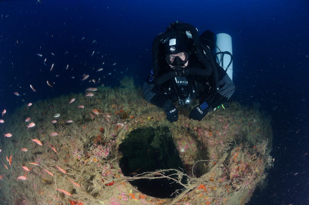 Subacqueo in assetto tecnico vicino alla torretta del sommergibile ricoperta di organismi marini e reti.