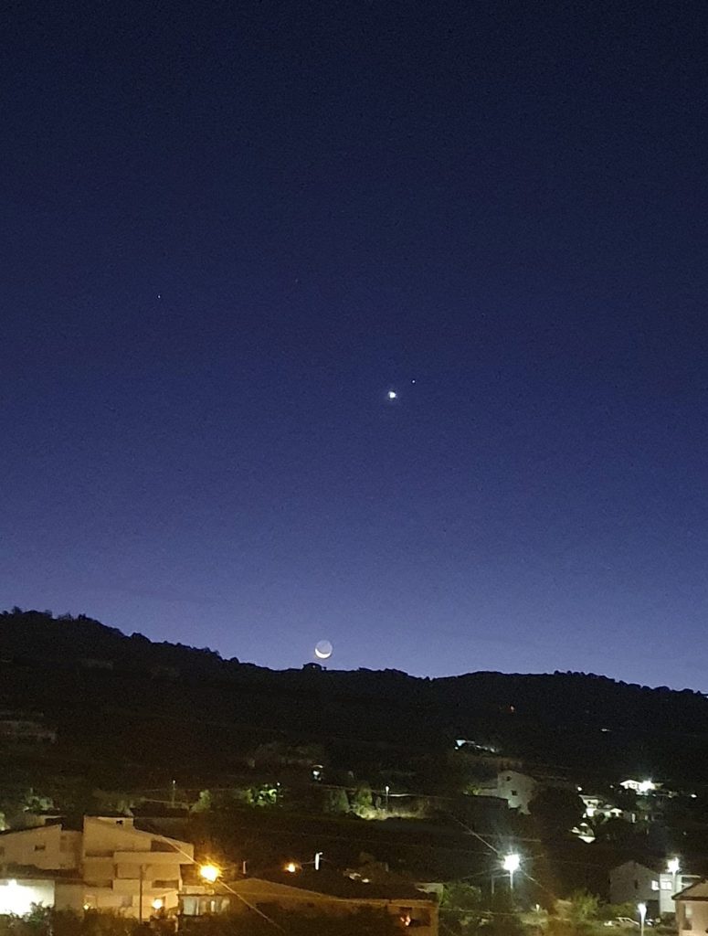 Congiunzione astrale tra la Luna, Venere e la stella Regolo ripresa all’alba sopra le colline del Mediterraneo, con il chiarore del giorno che inizia a illuminare il paesaggio.