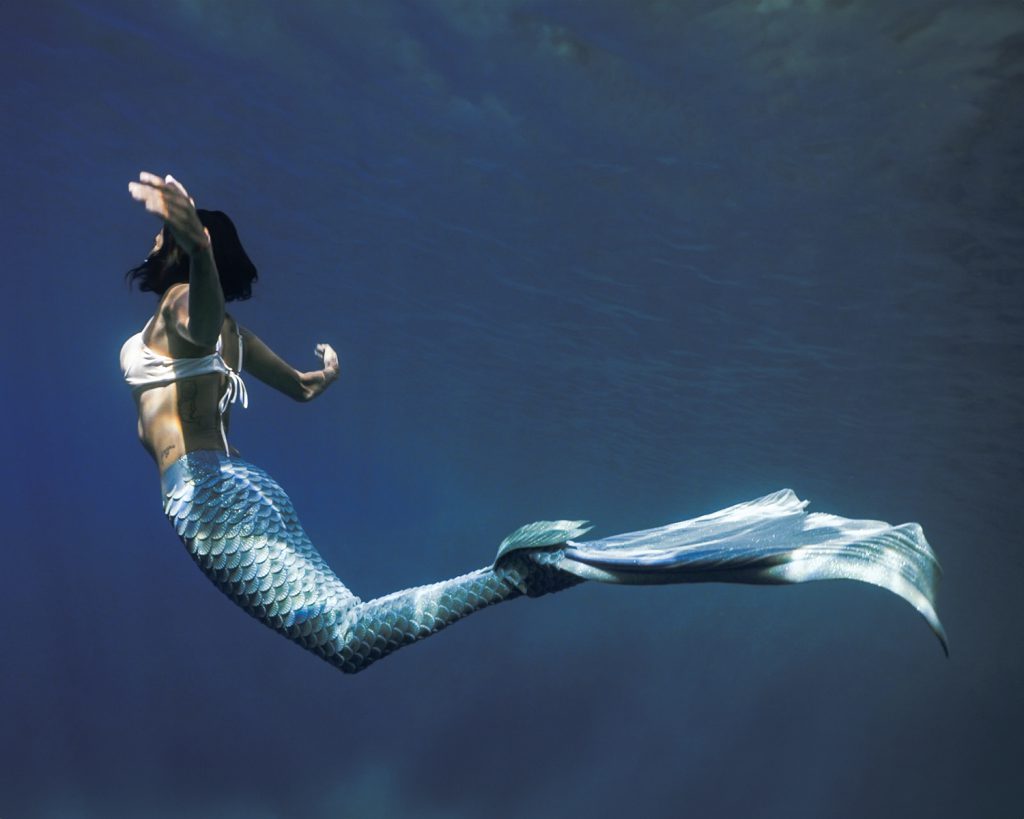 Ilaria Molinari con coda da sirena azzurra e squame, e top bianco, che nuota elegantemente sott'acqua.