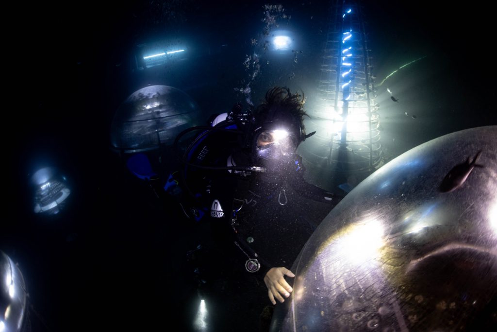 Subacqueo durante un’immersione notturna tra le biosfere del Giardino di Nemo a Noli, con illuminazione artificiale e strutture delle serre subacquee visibili sul fondale.